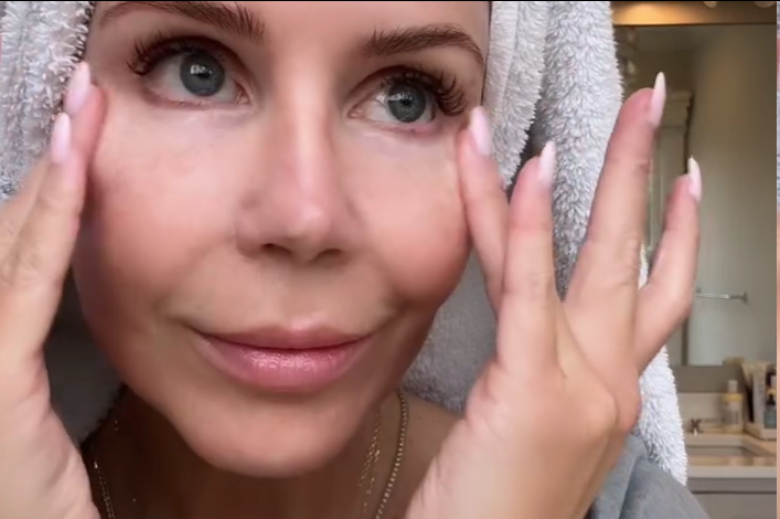 Close-up of a woman with a towel on her head, gently touching her face, showcasing clear skin and a subtle smile,